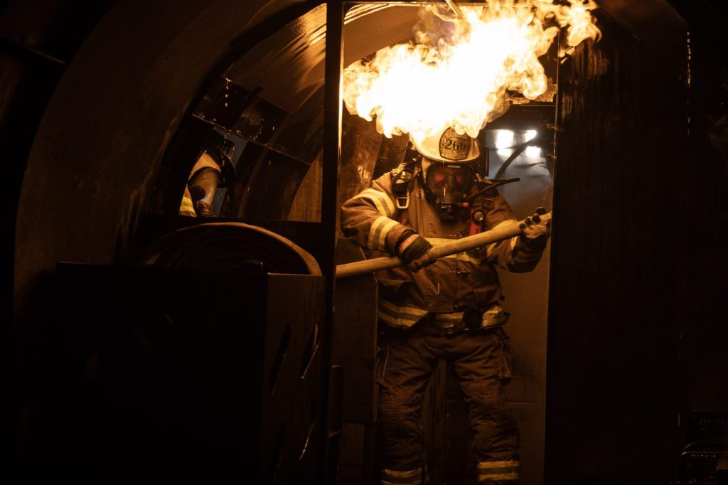 Firefighter with a hose line under rolling flames inside an aircraft cabin trainer
