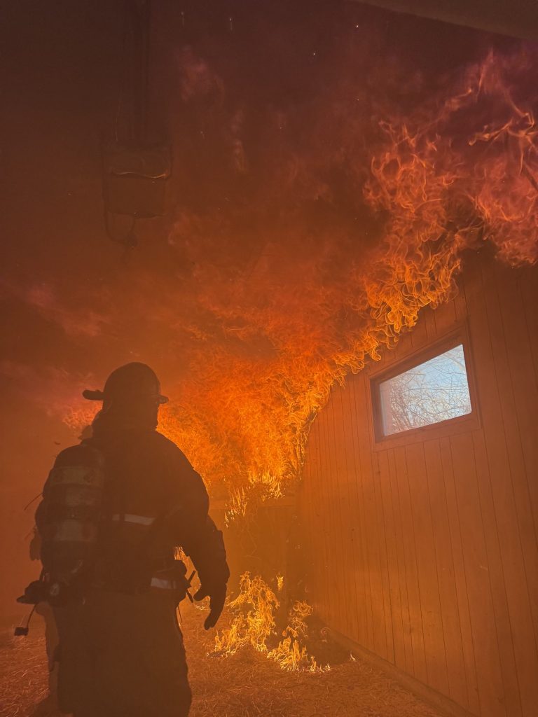 Firefighter in silhouette watching flames roll across a burn room ceiling