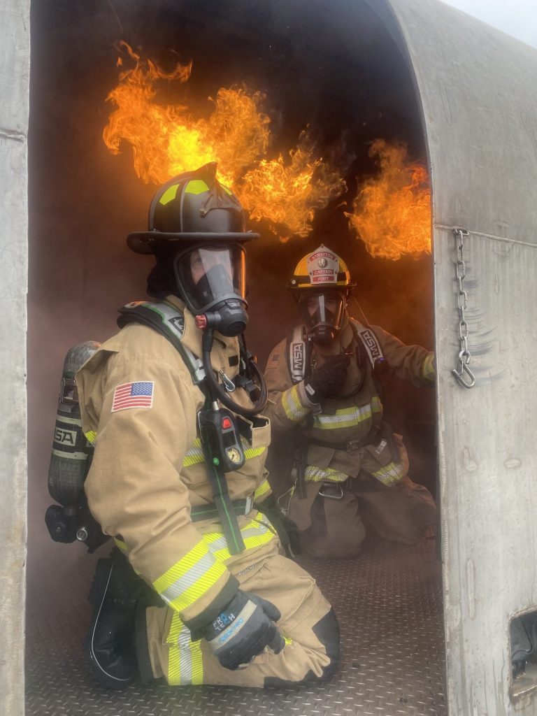Firefighters kneeling in an aircraft trainer doorway under flames