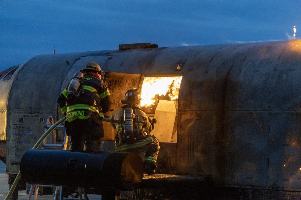 Firefighters advancing into a burning aircraft trainer at dusk