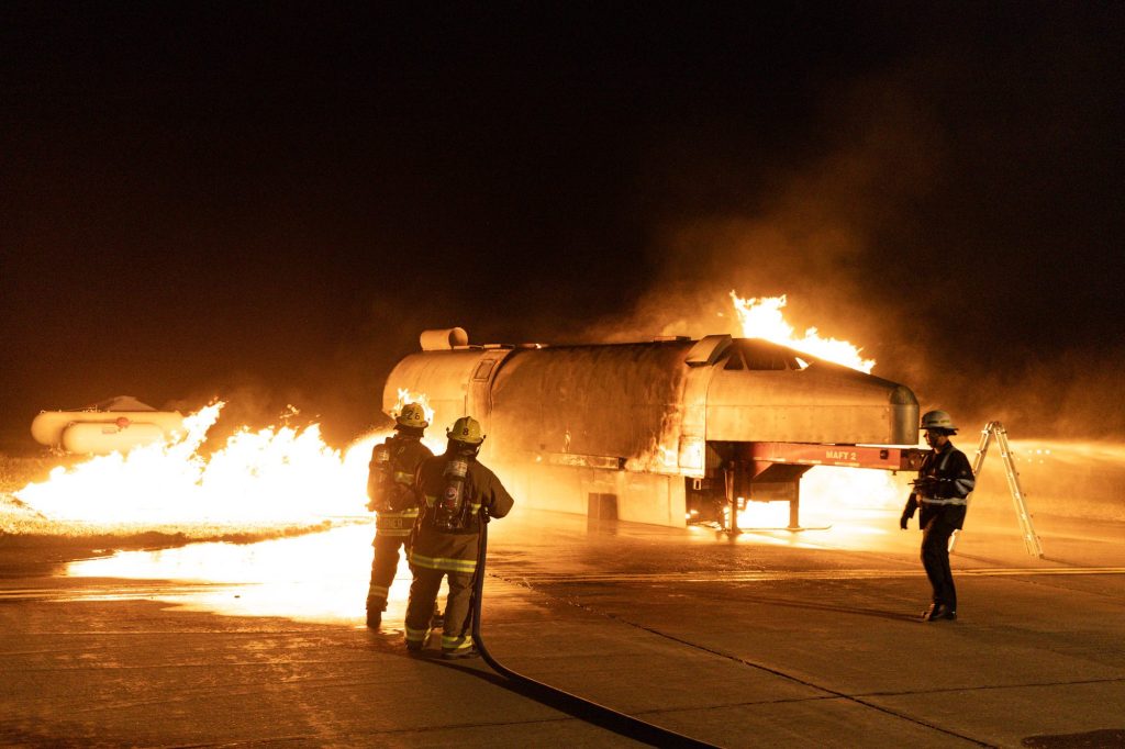 Hose team approaching an aircraft trainer with a ground fuel fire at night