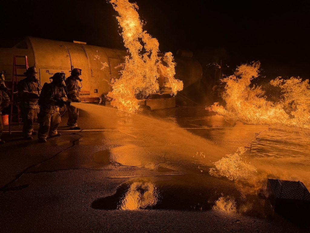 Hose teams attacking a large fuel fire beside an aircraft trainer at night