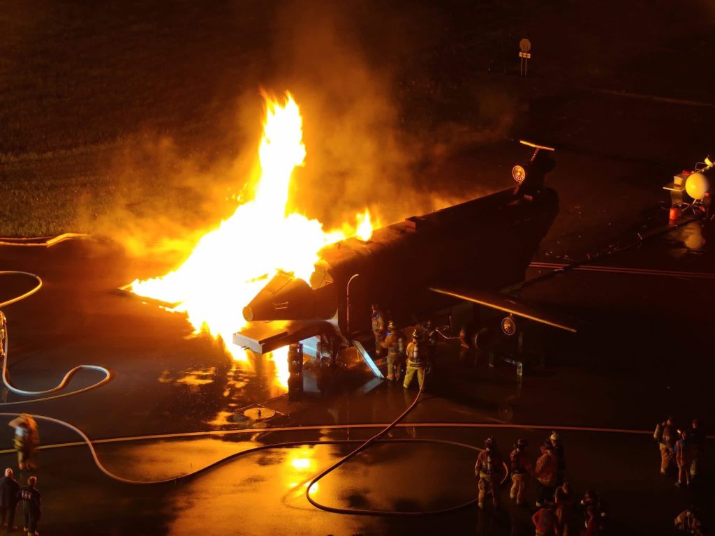 Elevated view of a fully involved aircraft fire trainer at night with hose teams staged nearby
