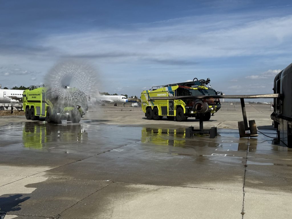 ARFF truck flowing its bumper turret beside a second crash truck near the aircraft fire trainer