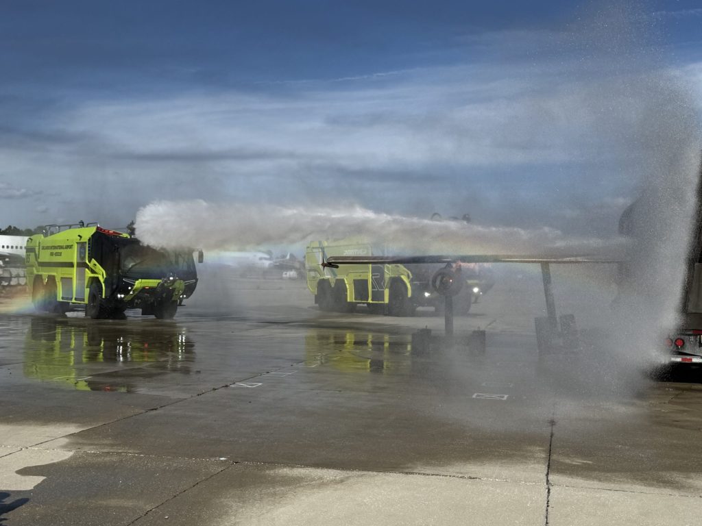 Two ARFF trucks flowing turrets toward the aircraft trainer wing with a rainbow in the spray