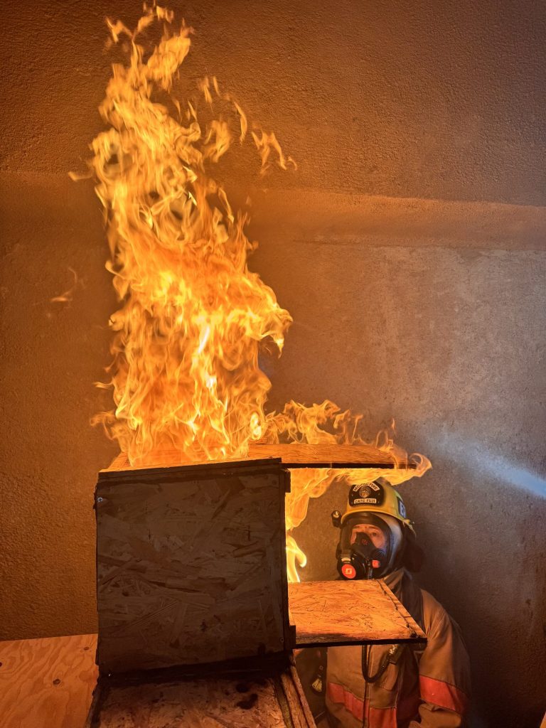 Flames rolling out of a fire behavior prop with a firefighter on air beside it