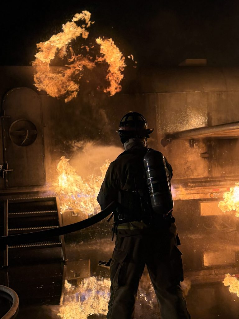 Firefighter in SCBA flowing a handline on fire at the aircraft trainer's cabin door at night