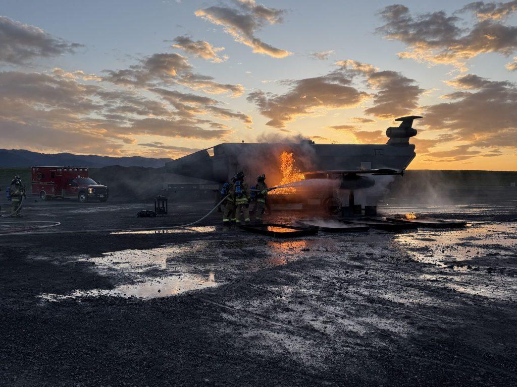Hose team attacking a fuselage fire on the aircraft trainer at sunset with an ambulance standing by